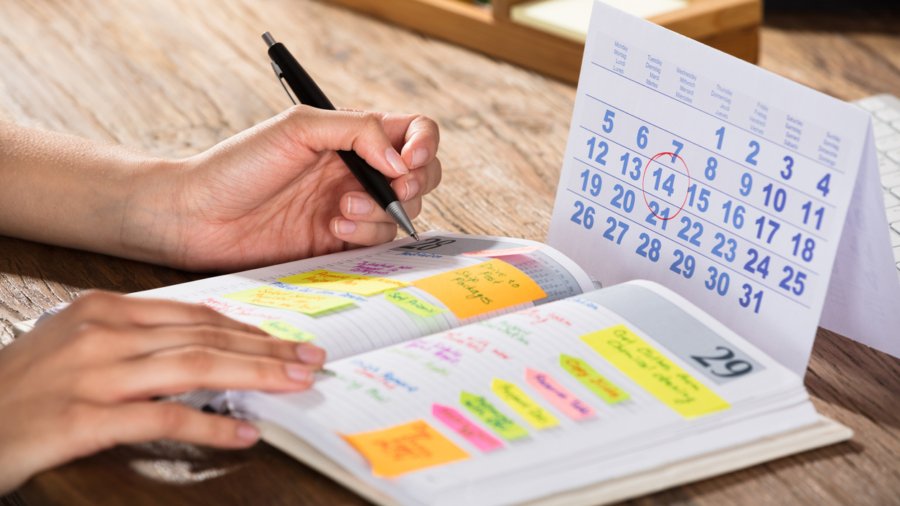 Close-up Of A Businessperson With Calendar Writing Schedule In Diary On Desk